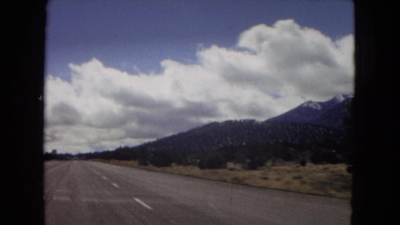 1967 Arizona road-trip view from inside a car, with an empty highway, distant mountain ridge, dry roadside terrain, and large cumulus clouds.