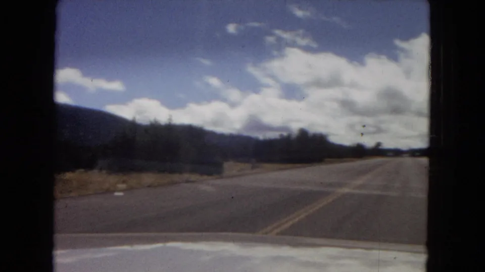 1967 Arizona home-movie driving frame showing a curving two-lane road, dry roadside landscape, distant hills, bright clouds, and the car hood at the lower edge.