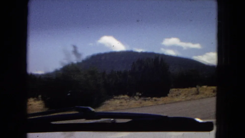 1967 Arizona car-window view with the windshield or dashboard edge in silhouette, roadside vegetation, a dark mountain ridge, and small white clouds against blue sky.