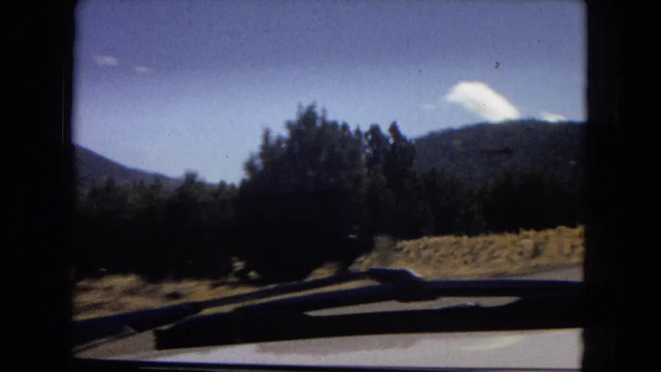 1967 8mm home-movie frame from a moving car, showing a windshield edge, roadside trees, dry ground, and distant mountains under a blue sky.