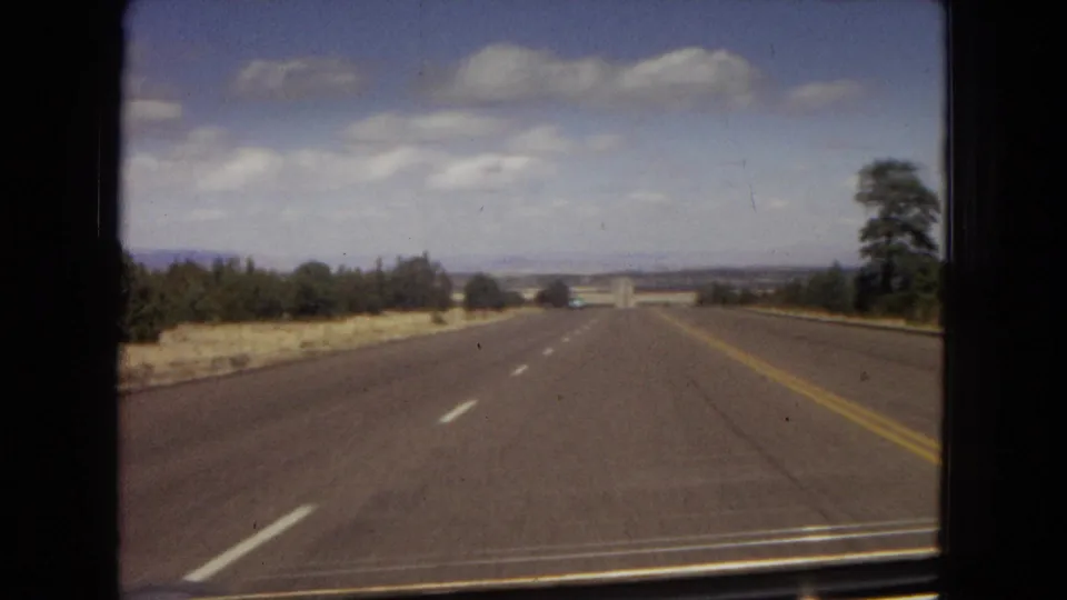 Moving-car view along an open Arizona highway in 1967, with lane markings, dry roadside vegetation, trees, distant landforms, and scattered clouds.