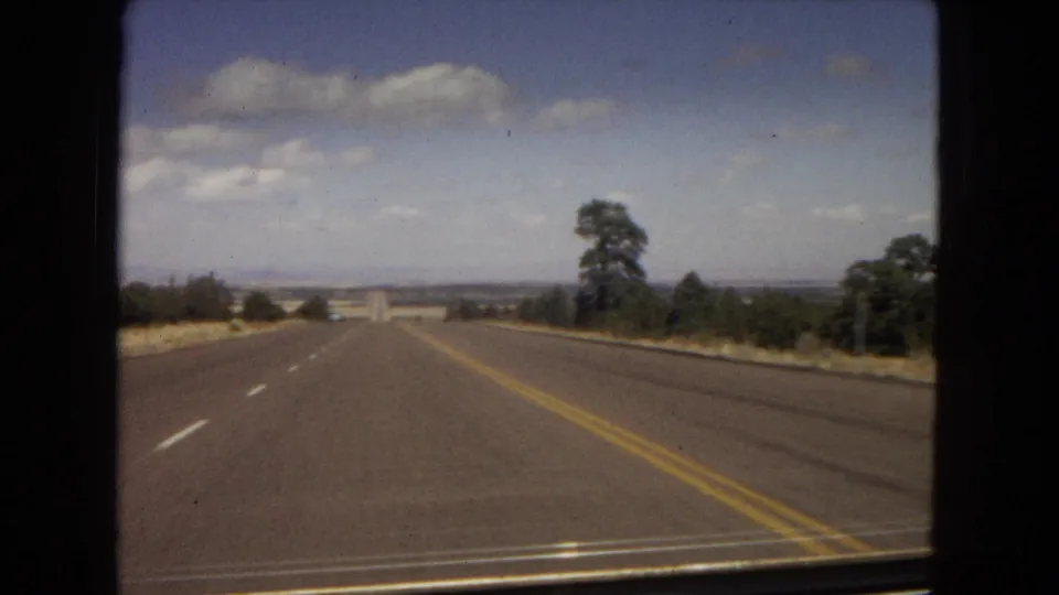 1967 home-movie road view looking forward along a two-lane highway, with dry roadside terrain, scattered trees, low distant hills, and a bright clouded sky.