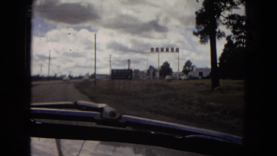 Moving-car frame near a roadside commercial area in 1967 Arizona, showing signs, utility poles, trees, low buildings, clouds, and a windshield wiper in the foreground.