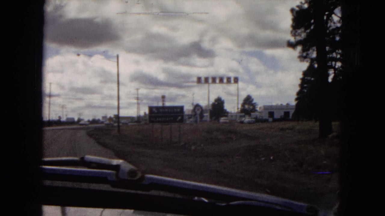 1967 Arizona 8mm road-trip frame showing a roadside approach through a vehicle window, with directional signs, utility poles, low buildings, and trees beneath heavy clouds.