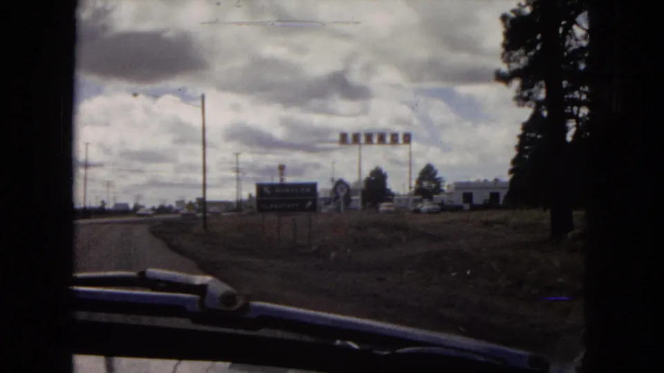 1967 Arizona 8mm road-trip frame showing a roadside approach through a vehicle window, with directional signs, utility poles, low buildings, and trees beneath heavy clouds.