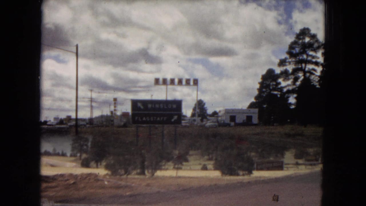 1967 Arizona road-trip frame with a prominent directional sign that appears to point toward Winslow and Flagstaff, seen beside a road with trees, buildings, and open sky.
