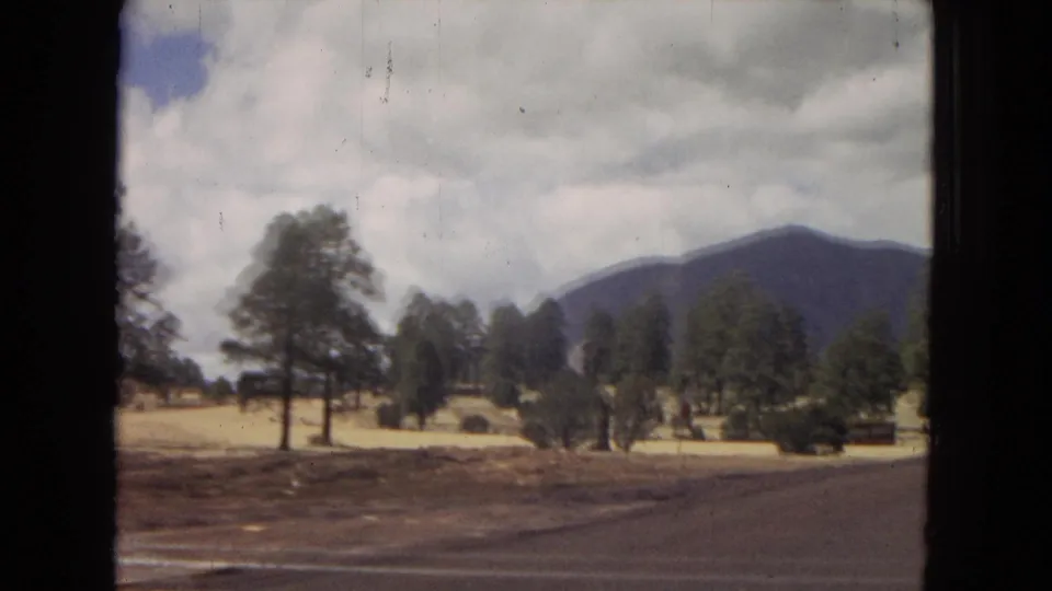 1967 Arizona 8mm road-trip frame showing roadside trees, open ground, a low sign, and a distant mountain under a broad cloudy sky.