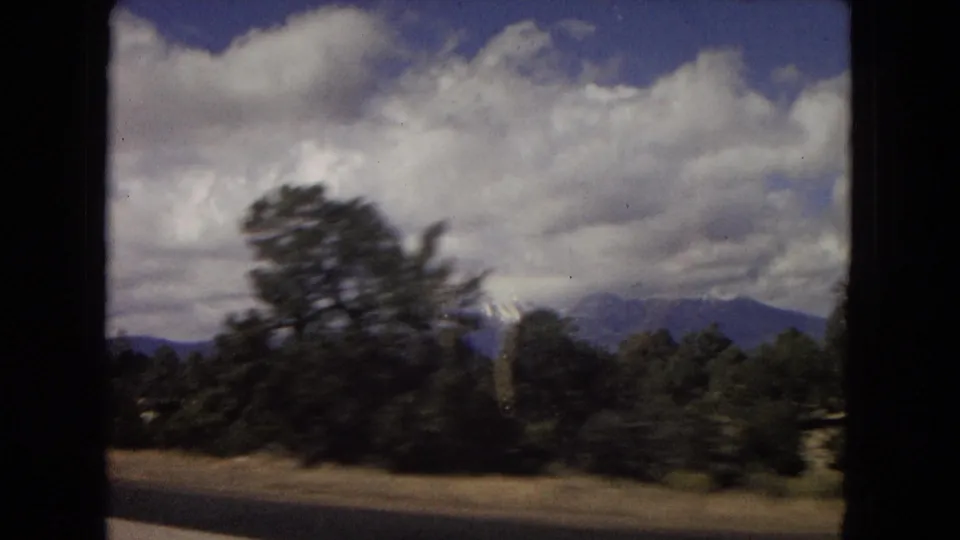 1967 Arizona 8mm road-trip frame with motion-blurred roadside trees, distant mountain forms, and dramatic clouds seen from a moving vehicle.