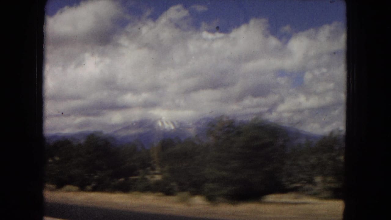 Arizona 1967 8mm frame showing roadside trees, low desert ground, distant mountains, and a cloudy sky filmed from a moving car.