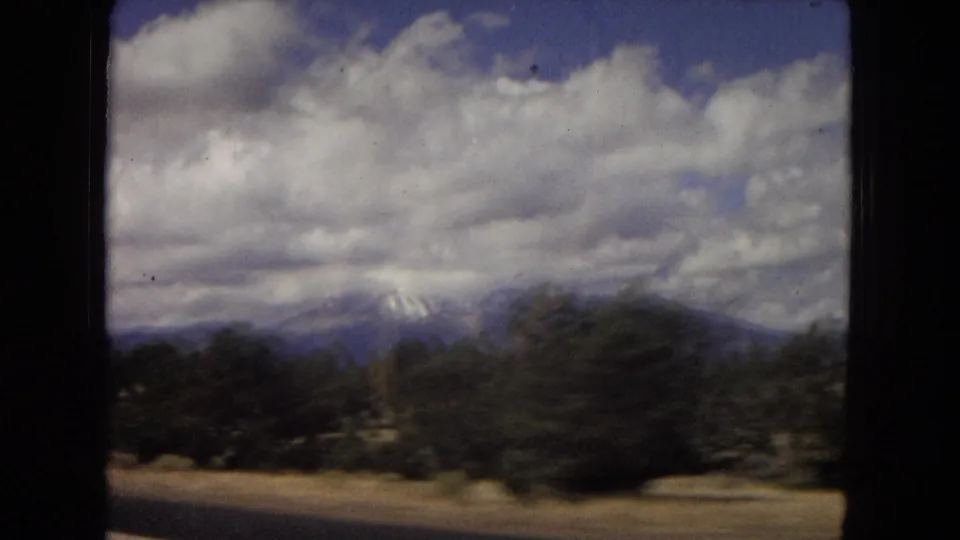 Arizona 1967 8mm frame showing roadside trees, low desert ground, distant mountains, and a cloudy sky filmed from a moving car.