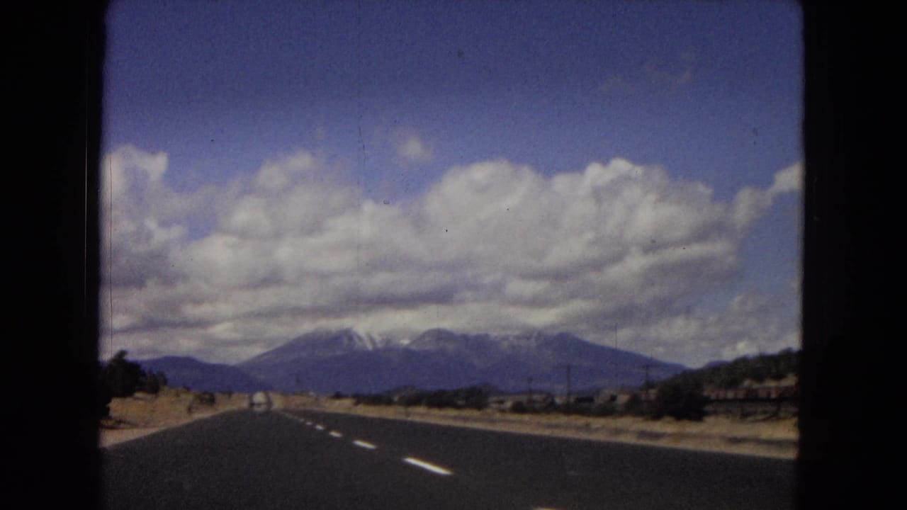 Arizona 1967 8mm frame with a paved highway stretching toward distant snow-capped mountains beneath a broad cloudy sky.