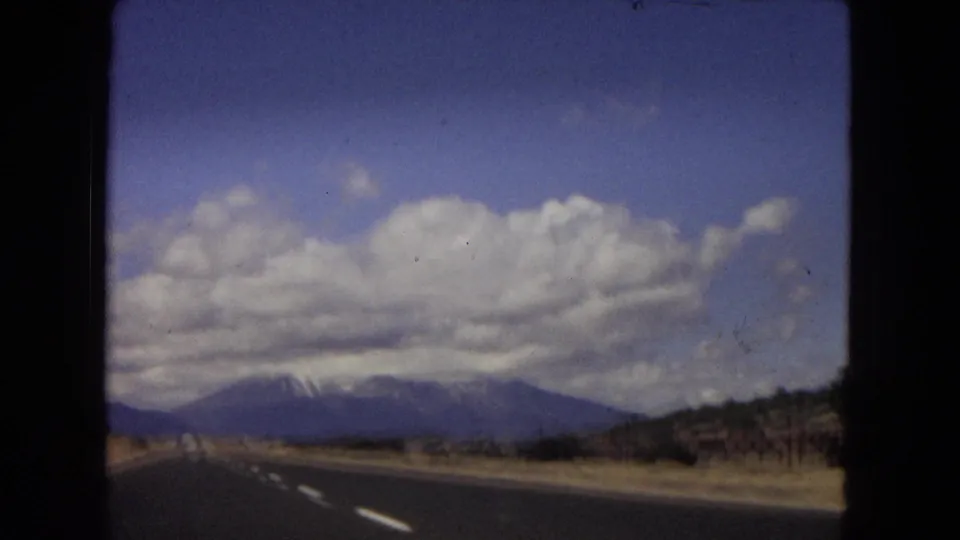 Arizona 1967 8mm frame showing a highway foreground, roadside vegetation, distant mountains, and large clouds from a moving vehicle perspective.