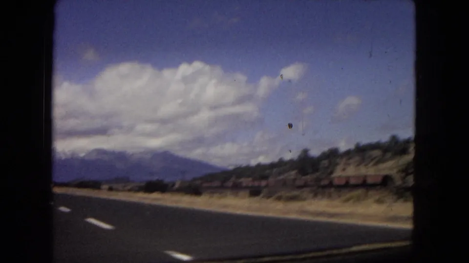 Arizona 1967 8mm frame with a highway edge, blurred roadside structures, trees, distant mountains, and cloud cover seen from a moving car.