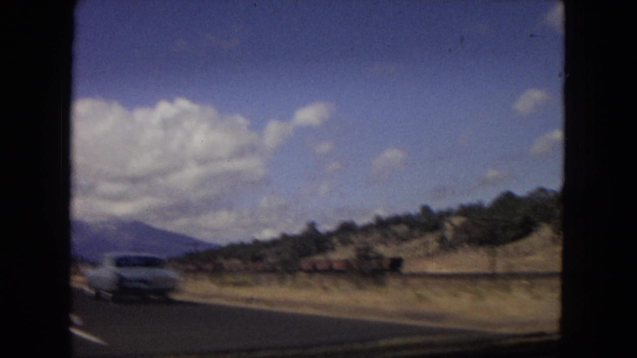 Metadata-supported 1967 Arizona road footage shows a light-colored automobile passing along a paved highway with desert hills, distant mountains, and a clouded blue sky.