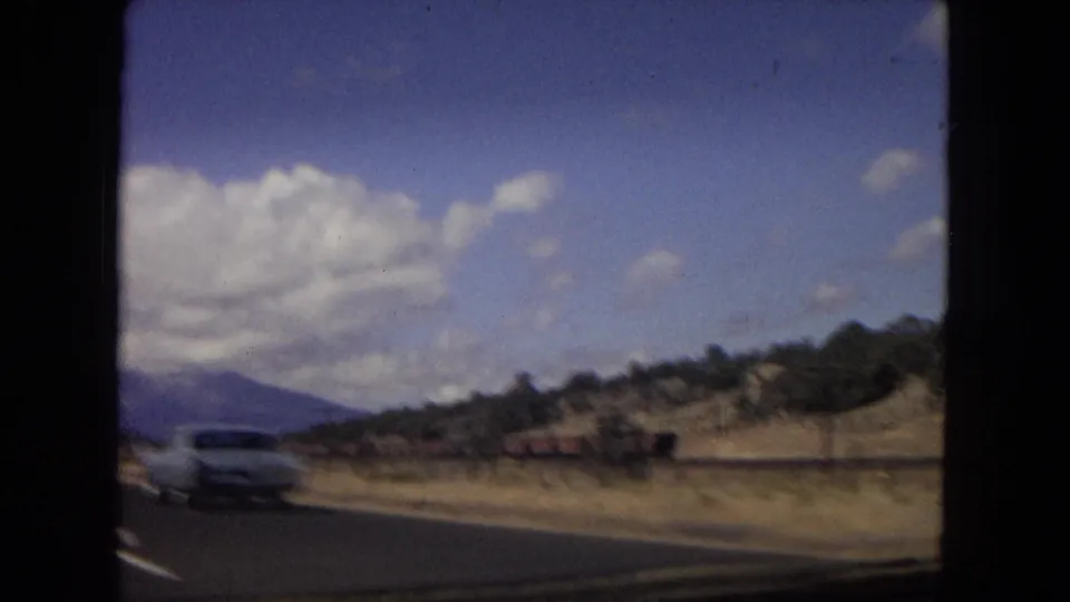 Metadata-supported 1967 Arizona road footage shows a light-colored automobile passing along a paved highway with desert hills, distant mountains, and a clouded blue sky.