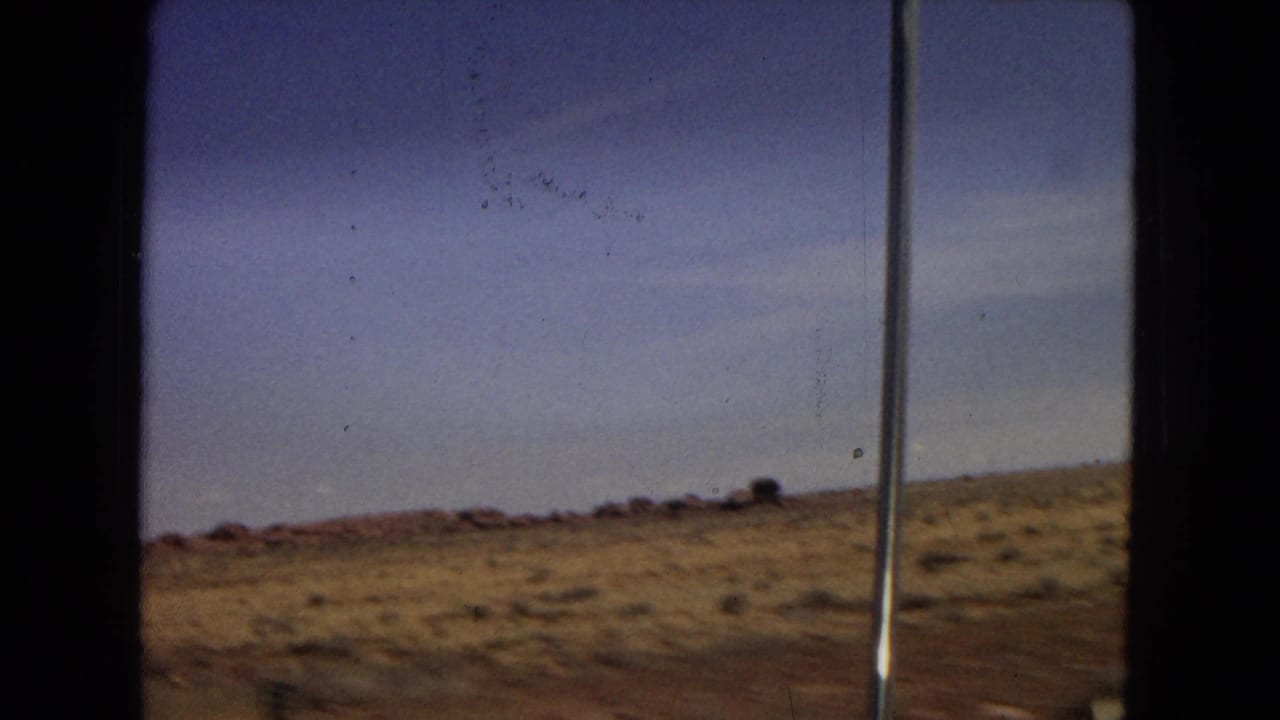 Metadata-supported Arizona highway footage shows open arid land, low scrub, reddish ground, a roadside pole, and a broad blue sky from a moving-car viewpoint.