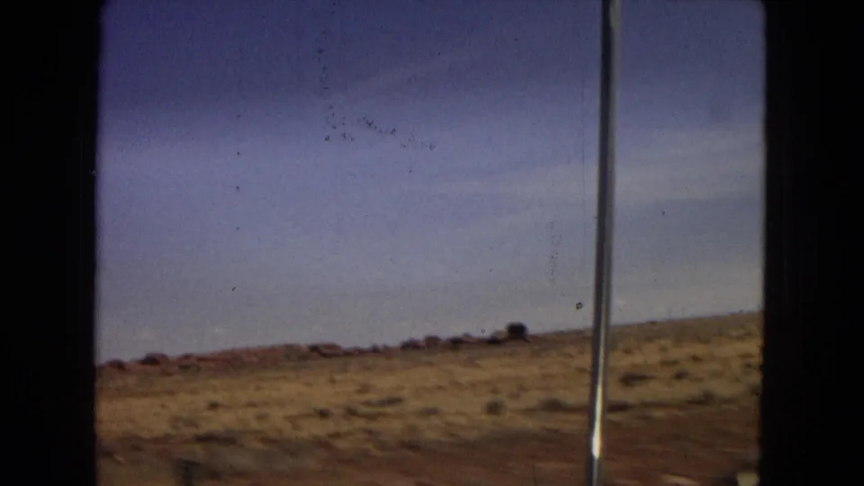 Metadata-supported Arizona highway footage shows open arid land, low scrub, reddish ground, a roadside pole, and a broad blue sky from a moving-car viewpoint.