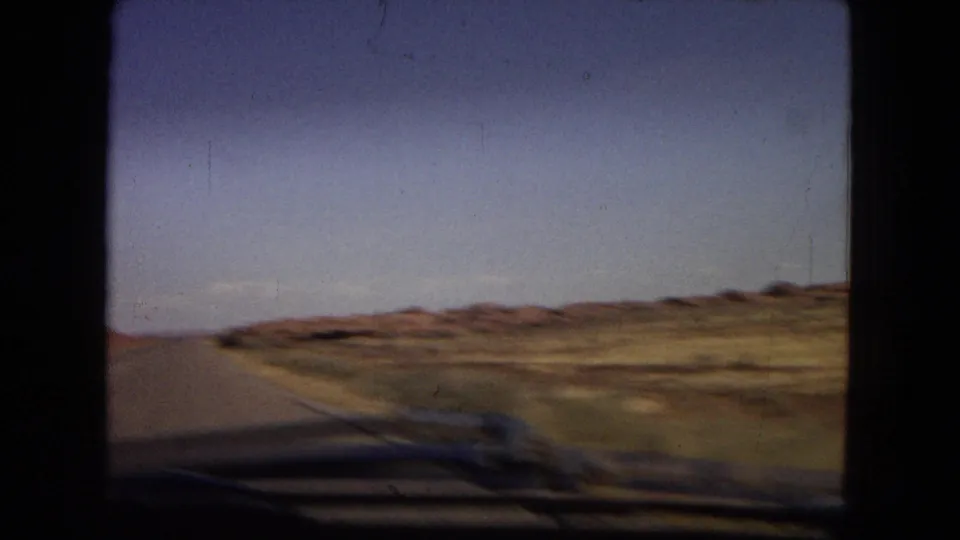 A moving-vehicle view in metadata-supported 1967 Arizona shows roadside desert terrain, low hills, blue sky, and the dark blurred edge of the car hood in the lower frame.