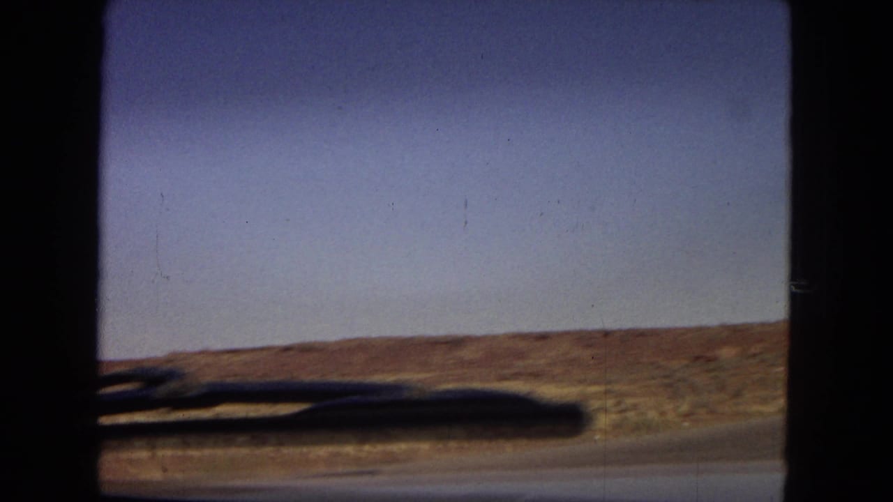 1967 Arizona 8mm frame showing a moving-car view across dry desert terrain beside an open highway under a clear blue sky.