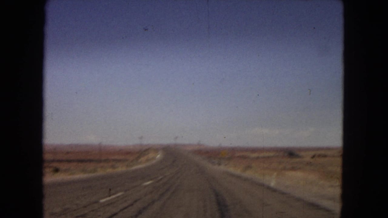 1967 Arizona 8mm frame looking forward down a long, nearly empty road through dry desert flats and distant low hills.