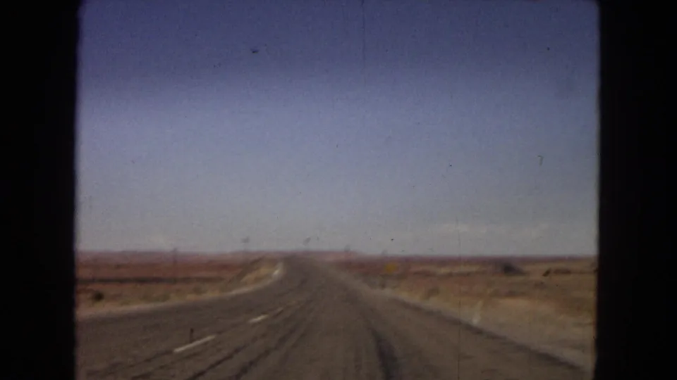 1967 Arizona 8mm frame looking forward down a long, nearly empty road through dry desert flats and distant low hills.