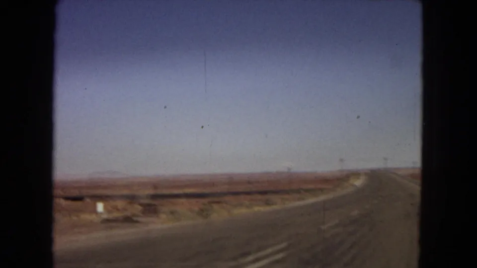 1967 Arizona 8mm frame from inside a moving vehicle, showing the road bending through open desert with a small roadside marker and distant horizon.
