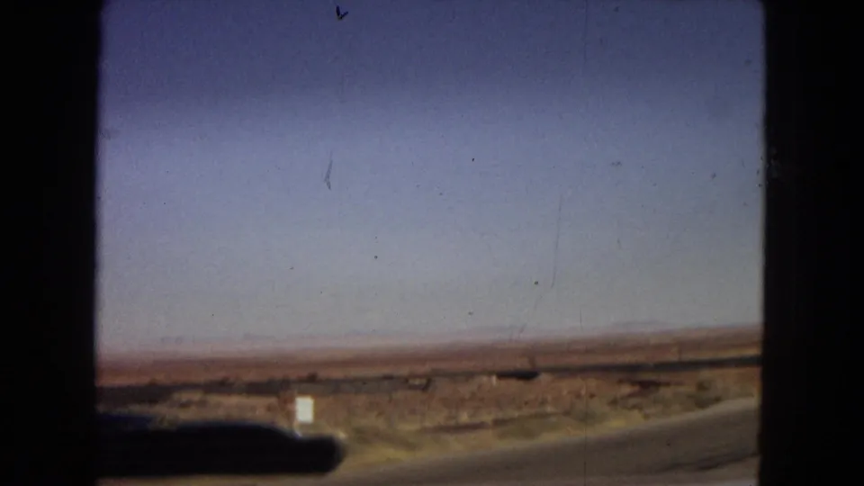 1967 Arizona 8mm frame showing a low desert horizon, road edge, and dark car hood from a windshield view during a scenic drive.