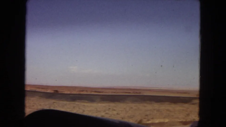 A low moving-vehicle view across dry roadside terrain toward a distant Arizona desert horizon in 1967 home-movie footage.