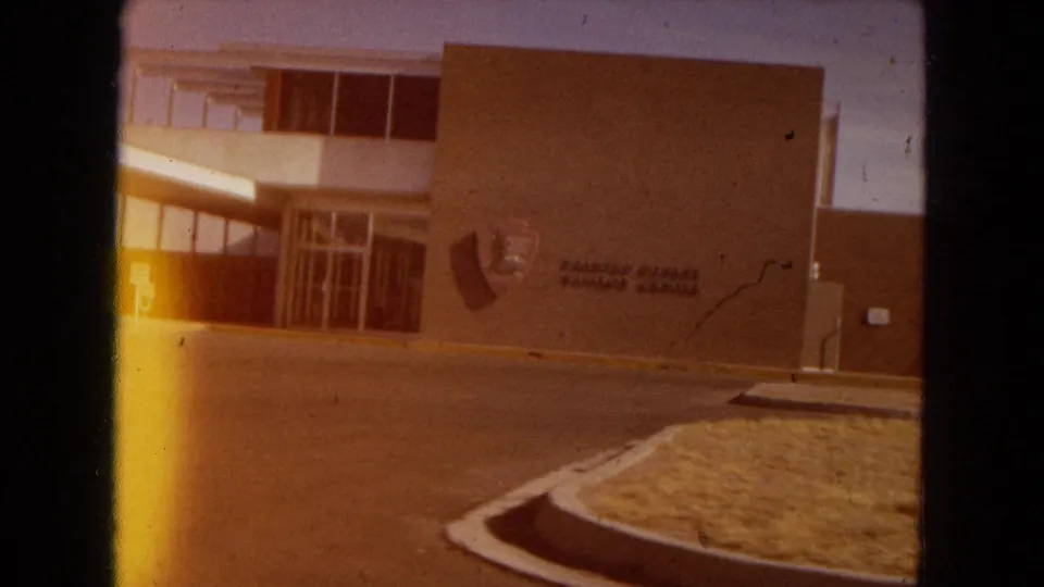 A brief 1967 Arizona roadside view of a modern institutional-looking building with exterior lettering, large windows, an entrance, curb, and dry landscaping.