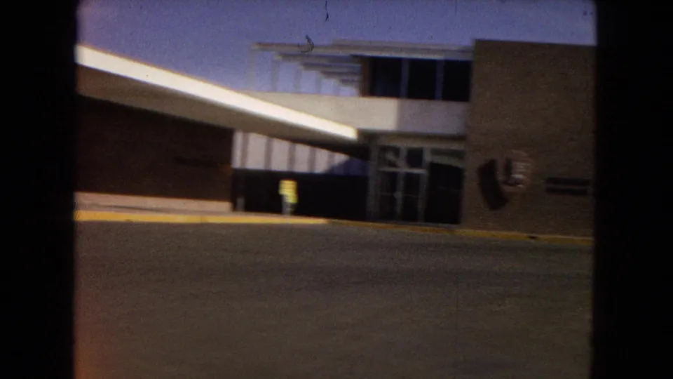 A 1967 United States home-movie frame shows a low mid-century school or institutional building with broad windows, a covered entrance, and a yellow curb in bright sun.