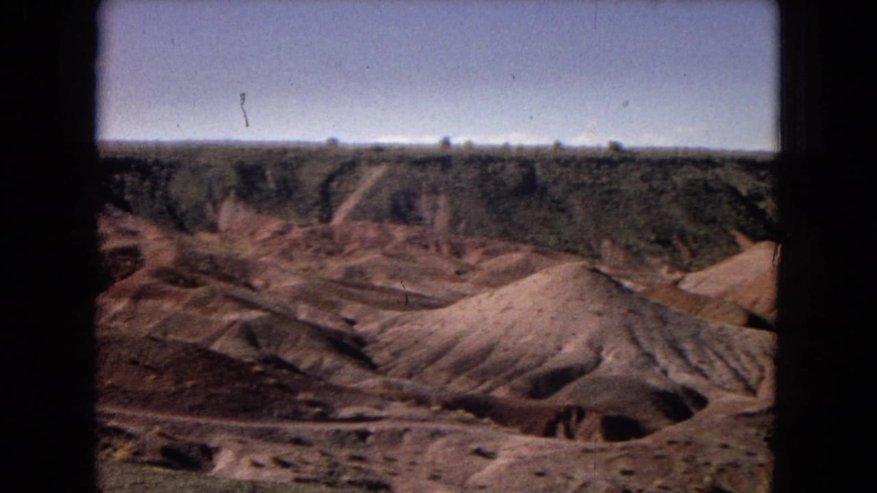 Arizona desert badlands appear in soft 8mm color, with pale eroded sand hills below a darker mesa edge and a clear blue sky.