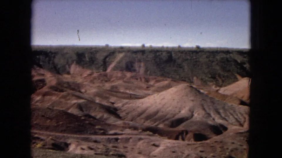 Arizona desert badlands appear in soft 8mm color, with pale eroded sand hills below a darker mesa edge and a clear blue sky.