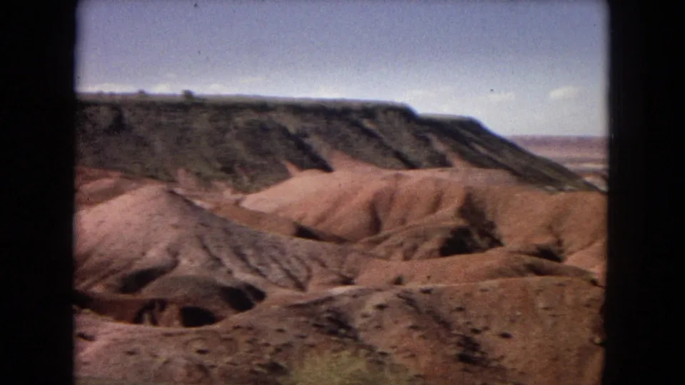 A wider Arizona desert view shows rounded pale sand hills, shadowed gullies, a dark mesa face, and distant flat terrain under a bright sky.