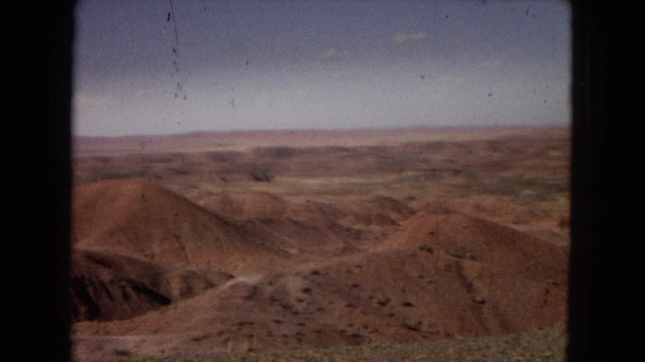 1967 Arizona 8mm frame showing reddish eroded desert hills, sparse scrub, distant horizon, and pale sky viewed through a dark central frame edge.