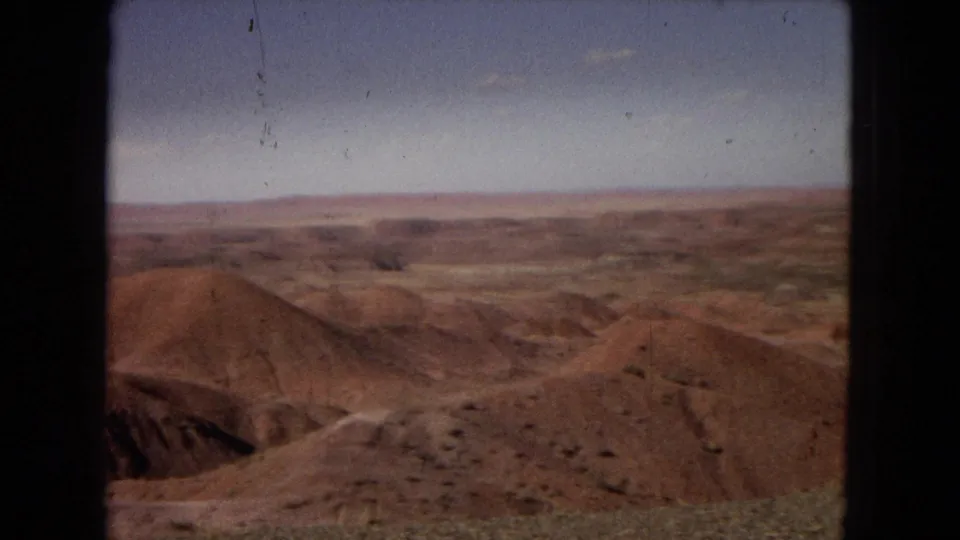 1967 Arizona 8mm frame showing reddish eroded desert hills, sparse scrub, distant horizon, and pale sky viewed through a dark central frame edge.