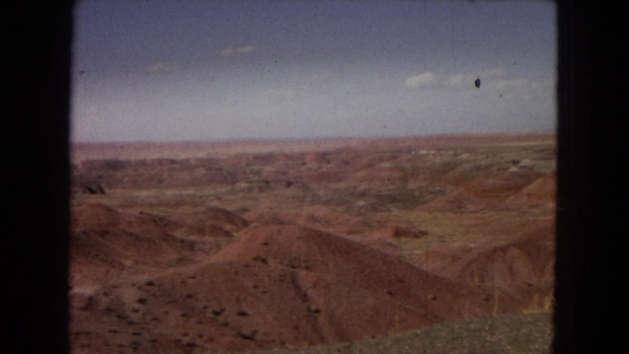 1967 Arizona 8mm view across red and brown desert formations with a low horizon, open sky, and dark film-frame borders visible at the edges.
