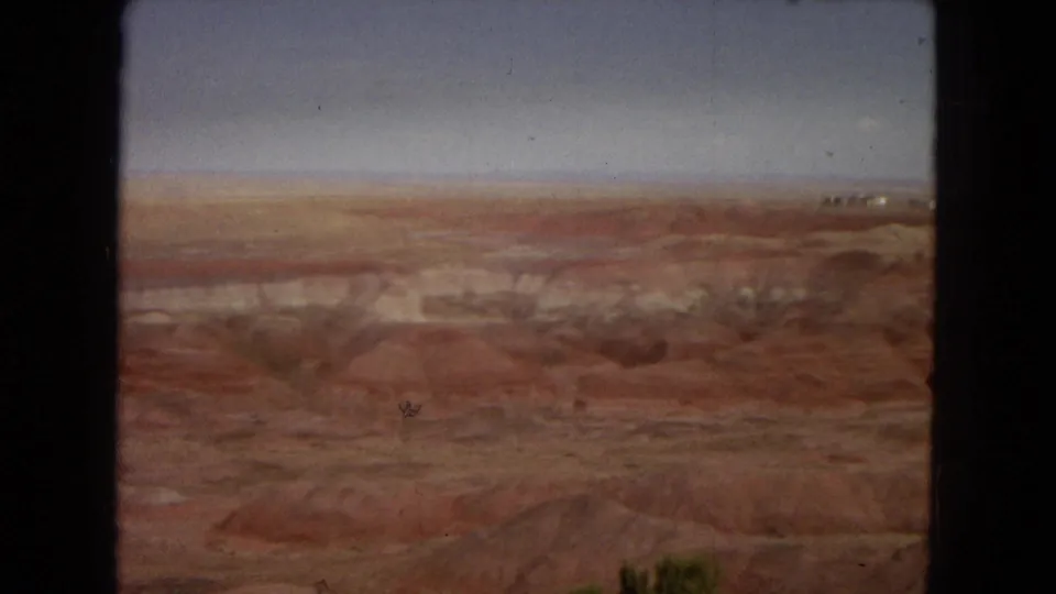 1967 Arizona 8mm frame with a soft panoramic view of desert flats, pale layered ridges, reddish foreground mounds, and a faint distant structure-like shape on the horizon.