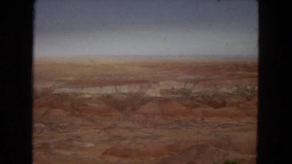 1967 Arizona 8mm frame showing a reddish desert basin, pale eroded bands, low scrub, and a hazy blue-gray sky within dark film-frame borders.