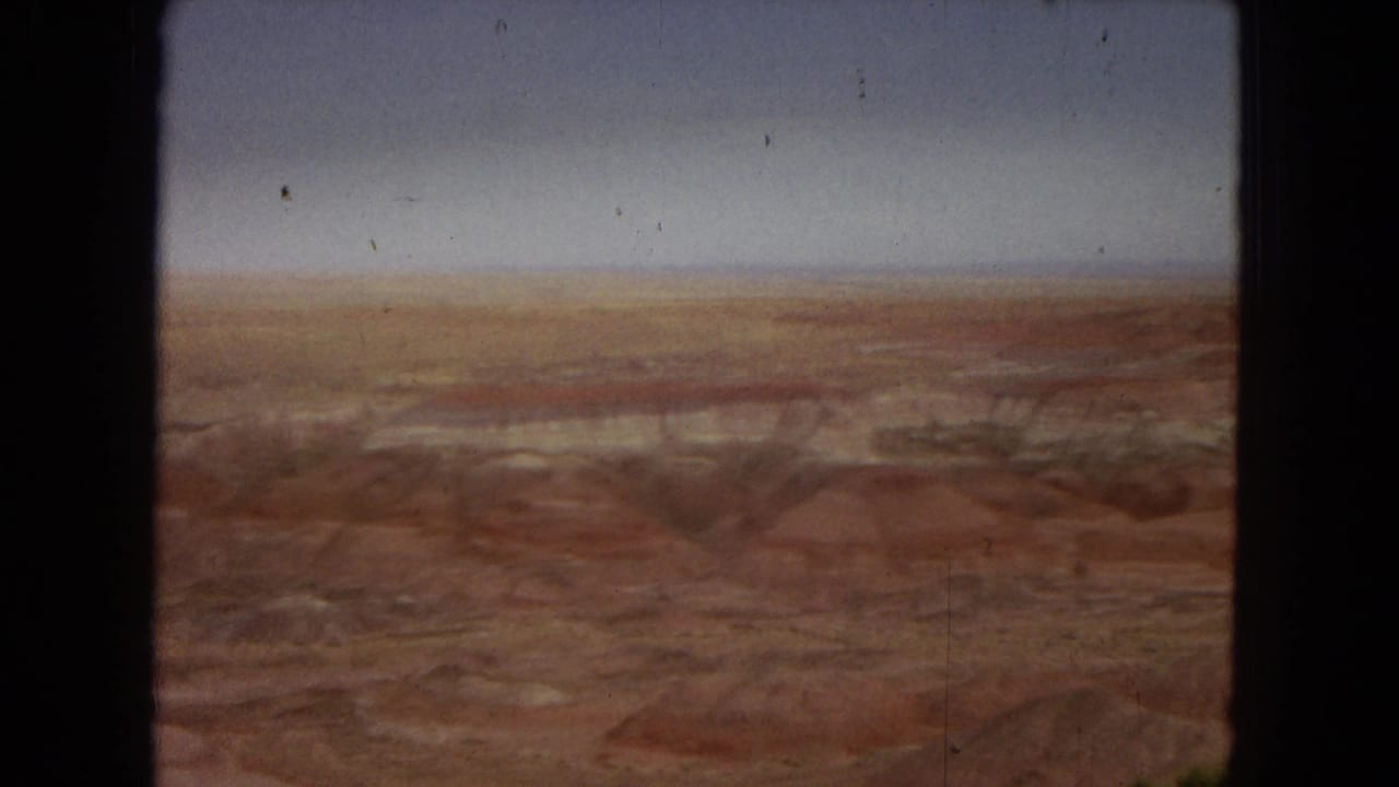 1967 8mm Arizona home-movie frame showing a wide red-brown desert or canyon landscape under a hazy sky, with dark gate borders and visible film wear.