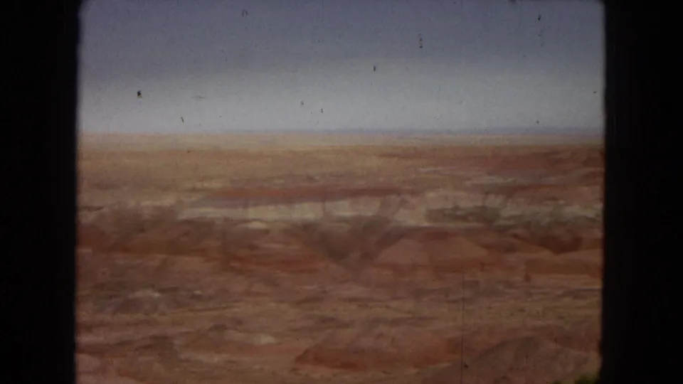 1967 8mm Arizona home-movie frame showing a wide red-brown desert or canyon landscape under a hazy sky, with dark gate borders and visible film wear.