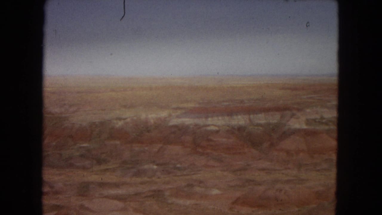 1967 8mm Arizona landscape frame with distant reddish and brown strata stretching across barren canyon-like terrain below a pale, hazy sky.
