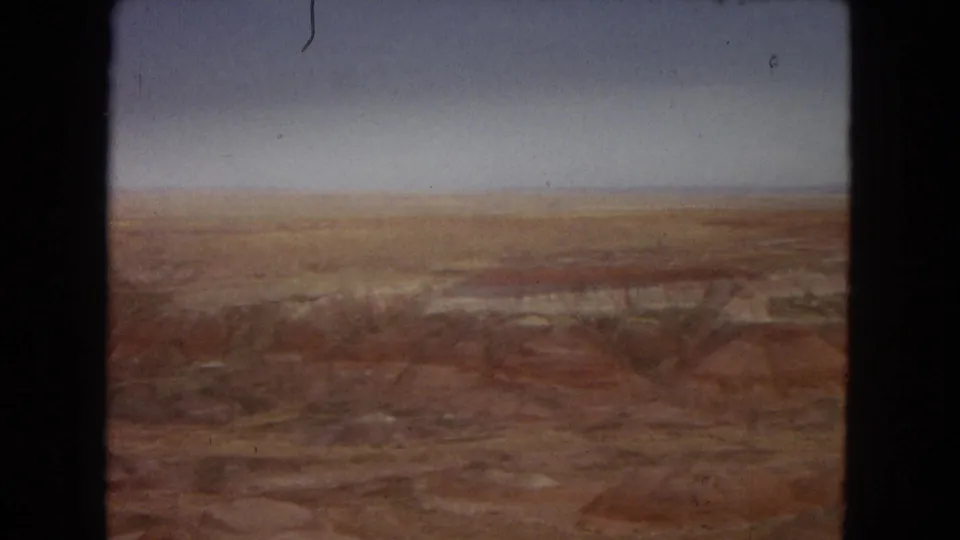 1967 8mm Arizona landscape frame with distant reddish and brown strata stretching across barren canyon-like terrain below a pale, hazy sky.