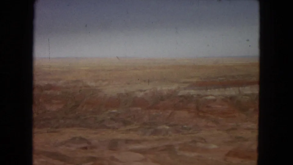 1967 8mm Arizona home-movie still showing a soft, wide view of barren red-brown desert terrain and a low distant horizon with visible grain and gate shadow.