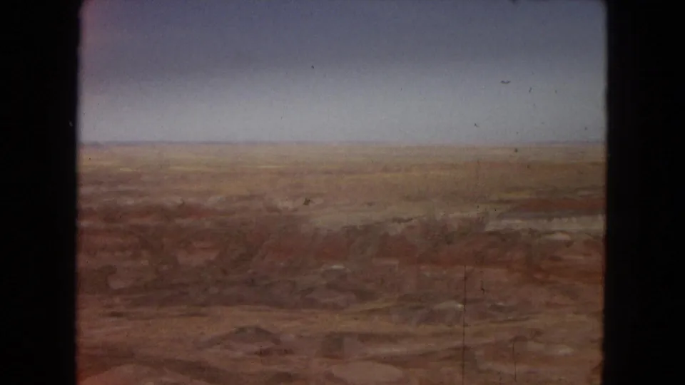 Final 1967 8mm Arizona frame holding on a distant arid canyon-like landscape, with muted red-brown layers, pale horizon, and visible film scratches.