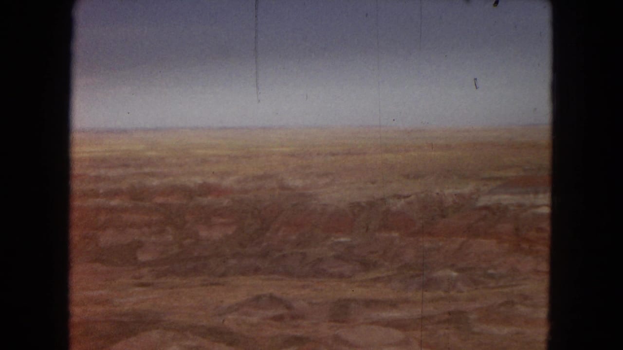 1967 Arizona home-movie frame showing a broad elevated view across red and ochre desert badlands beneath a muted blue sky.