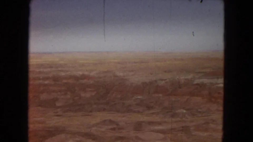 1967 Arizona home-movie frame showing a broad elevated view across red and ochre desert badlands beneath a muted blue sky.