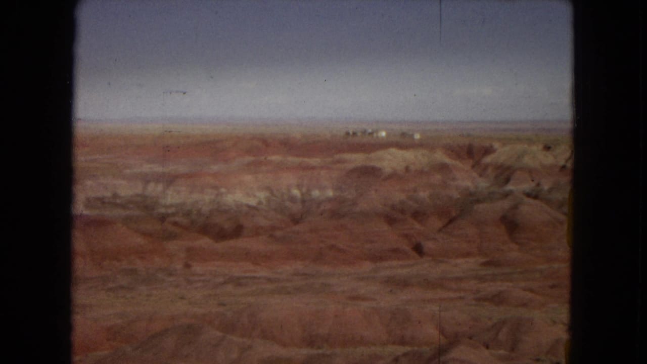 Elevated Painted Desert view with eroded red-brown formations, lighter sediment bands, and a flat distant horizon in 8mm color.