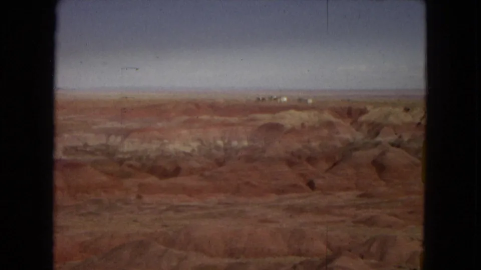 Elevated Painted Desert view with eroded red-brown formations, lighter sediment bands, and a flat distant horizon in 8mm color.