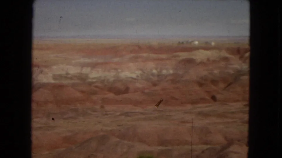 Closer 8mm frame of Arizona desert terrain, showing reddish slopes, pale stratified bands, and uneven eroded channels.