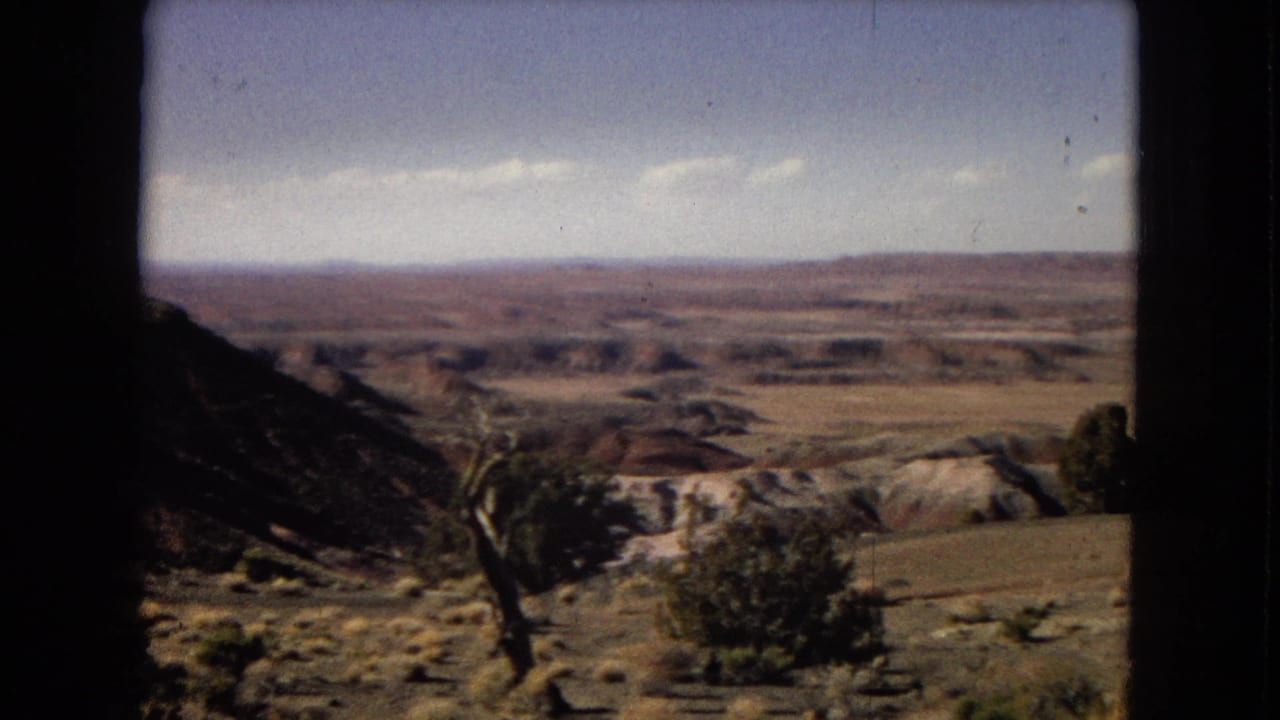 1967 Arizona 8mm frame with a broad desert horizon, pale rock bands, scattered vegetation, and a dark vertical edge suggesting filming through a vehicle or window opening.